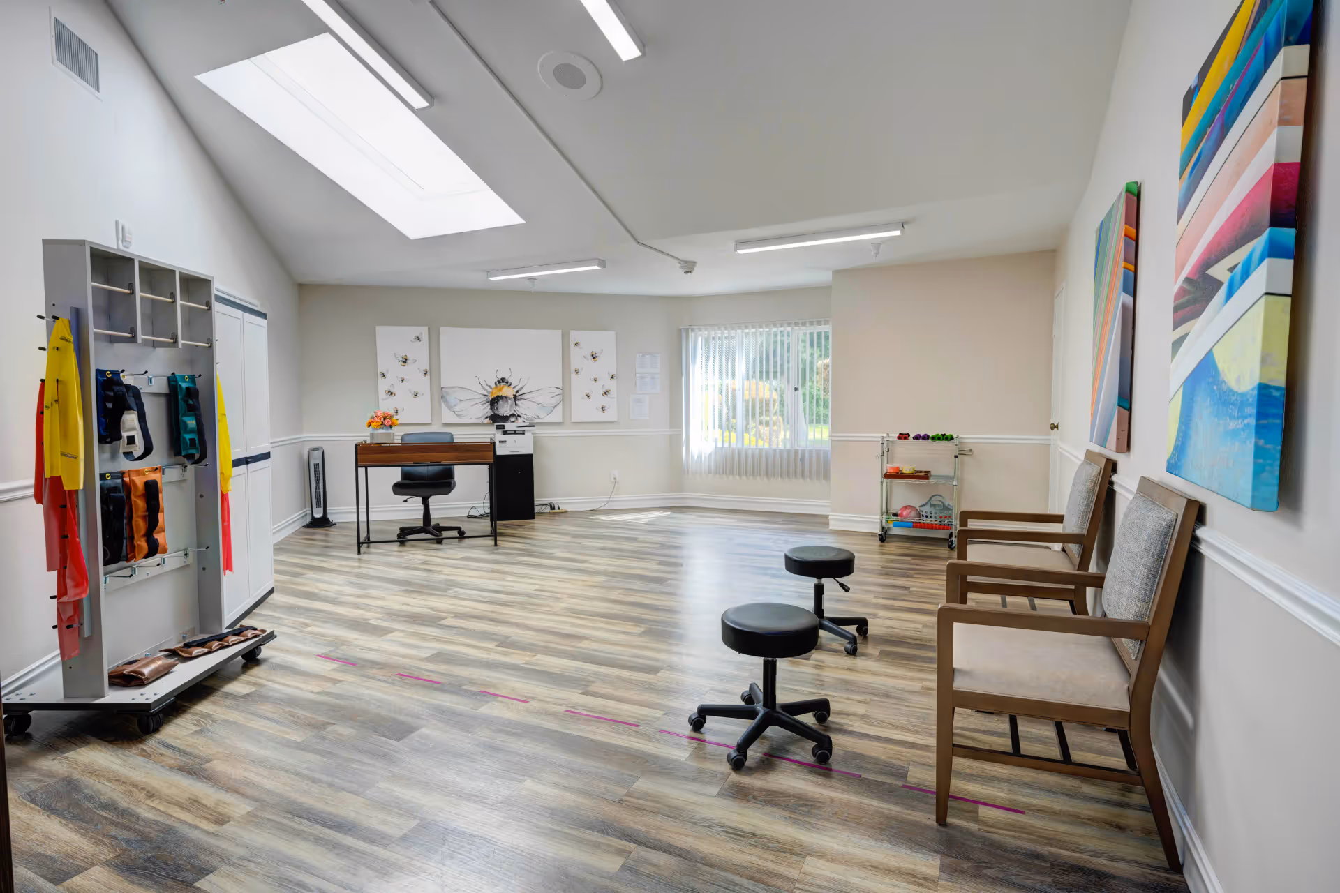 A spacious, well-lit room with wood-patterned flooring and a skylight. The room contains two wooden chairs with cushions along one wall, two black rolling stools in the center, and a small desk with a chair against the far wall. Colorful exercise bands hang on a rack to the left, and a small cart with various exercise equipment is near a window with vertical blinds. The walls are decorated with large bee-themed artwork and colorful abstract paintings.