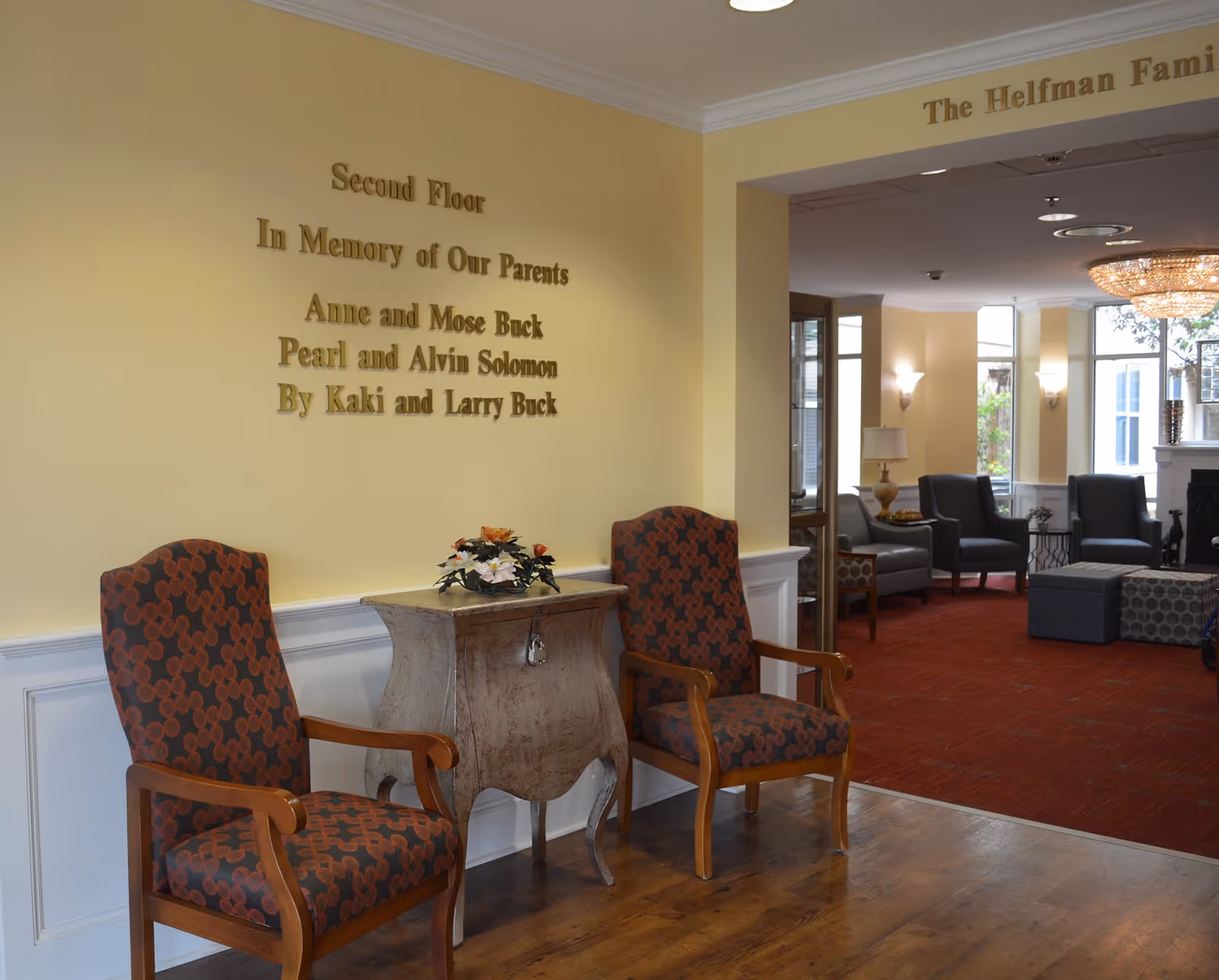 A sitting area inside The Medallion Jewish Assisted Living with two patterned armchairs flanking a small wooden table with a floral arrangement. The wall behind the chairs has a memorial plaque dedicated to Anne and Mose Buck, Pearl and Alvin Solomon by Kaki and Larry Buck. In the background, there is a larger lounge area with additional seating, a chandelier, and large windows letting in natural light.