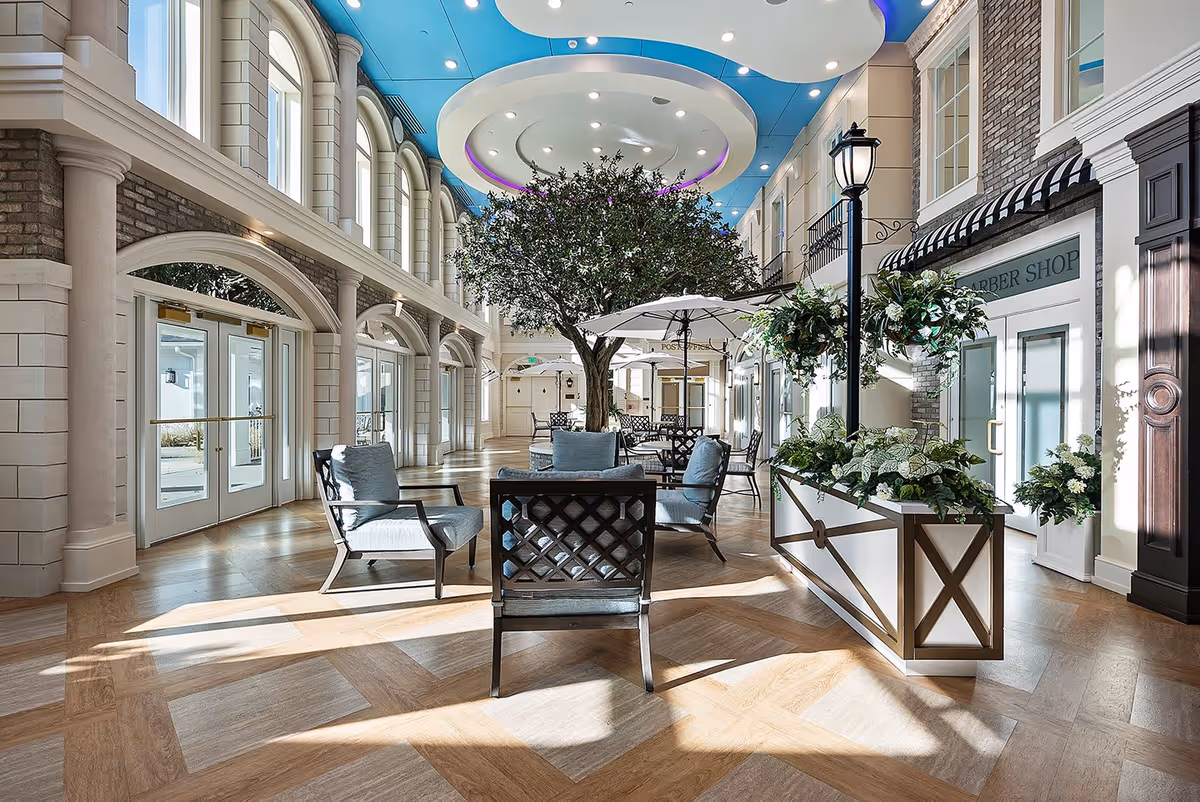 A bright and spacious indoor common area with large arched windows letting in natural light. The room features a blue ceiling with circular recessed lighting, a large artificial tree in the center, comfortable seating with cushioned chairs, umbrellas over tables, decorative plants, and a streetlamp-style light fixture. There is a sign for a barber shop on one side and a post office in the background.