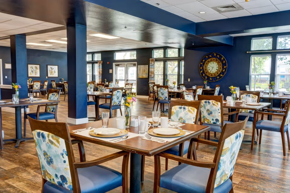 A bright and spacious dining room with multiple wooden tables and chairs featuring floral-patterned upholstery. Each table is set with plates, cups, glasses, and silverware, and decorated with small flower vases. Large windows allow natural light to fill the room, and a large decorative clock is mounted on a dark blue wall.