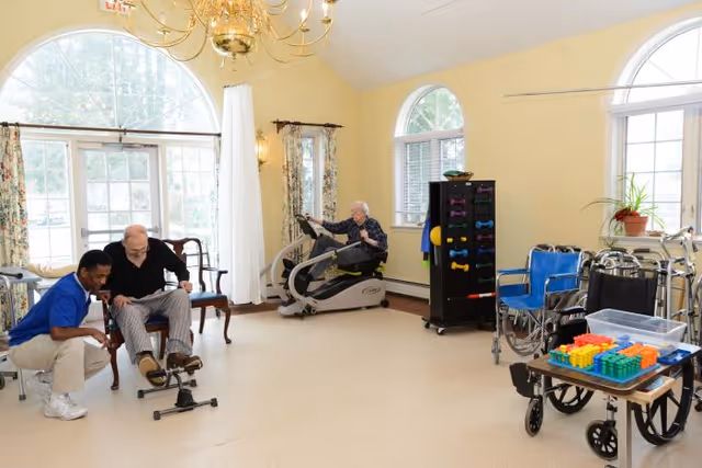 A senior man using a seated exercise machine in a bright room with large arched windows. Another senior man is seated on a chair while a caregiver assists him with a foot pedal exerciser. The room contains exercise equipment, a wheelchair, walkers, a table with colorful blocks, and a rack with small dumbbells. The walls are painted light yellow and there is a chandelier hanging from the ceiling.