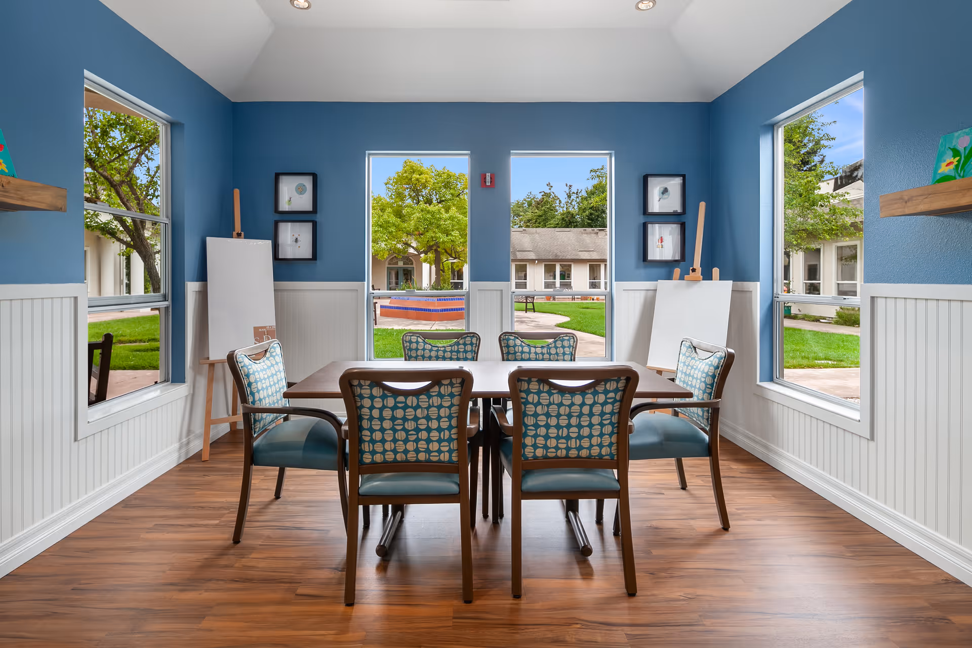 Bright blue-walled activity room with a table and six patterned chairs facing windows overlooking a courtyard.