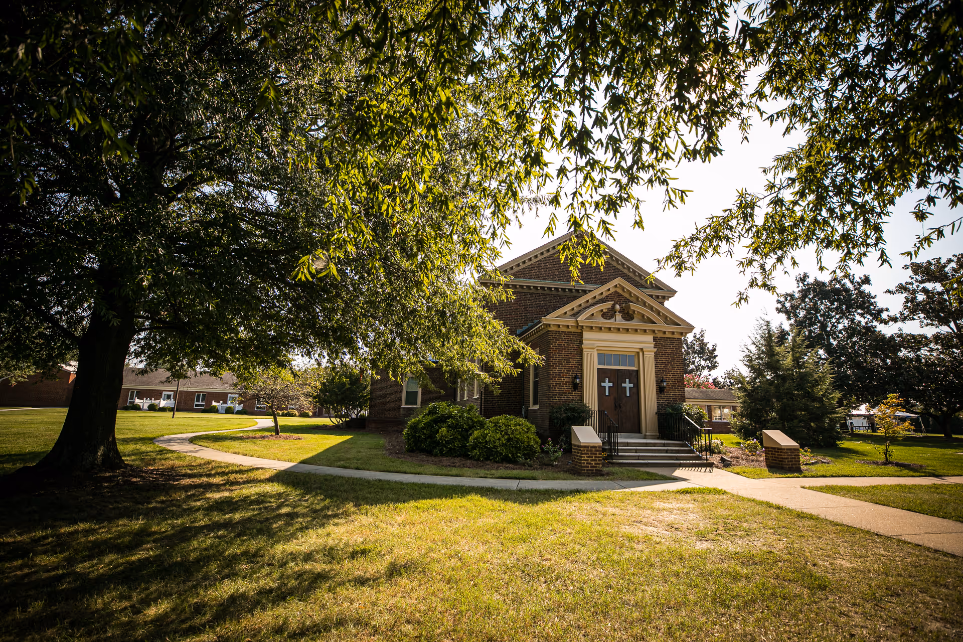 A small brick chapel building with two white crosses on the wooden double doors, surrounded by green grass, trees, and shrubs under a sunny sky.
