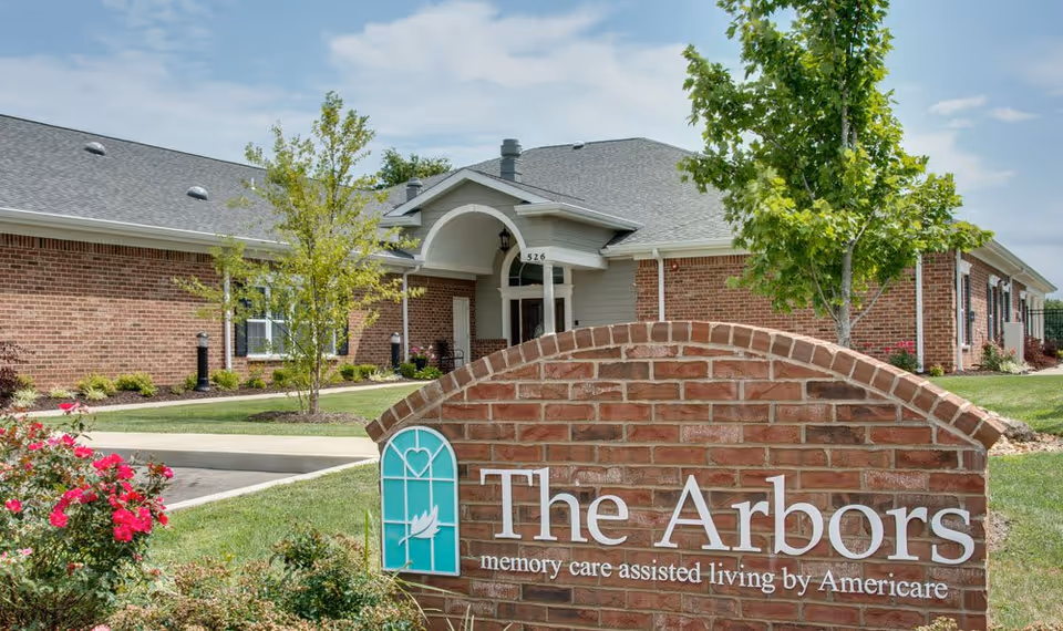 Exterior view of a single-story brick building with a gray roof, surrounded by green grass, small trees, and flowering bushes. In the foreground, there is a brick sign that reads 'The Arbors memory care assisted living by Americare'.