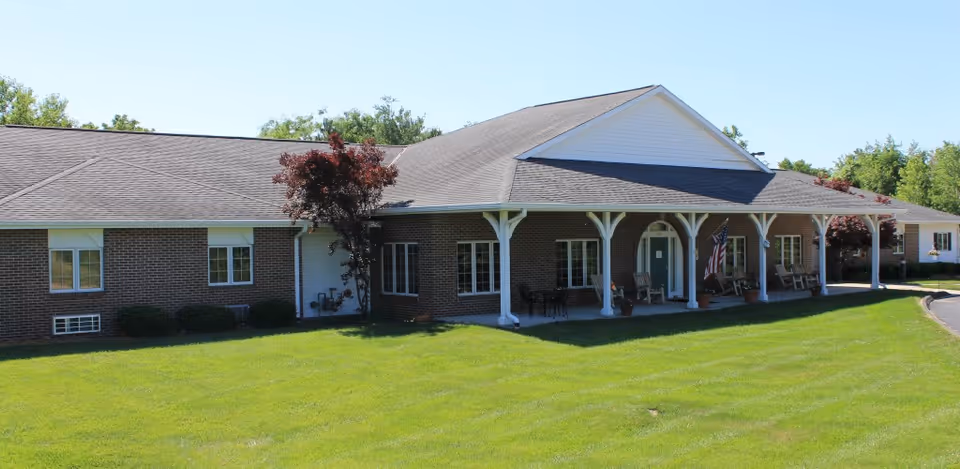 Front exterior of a single-story brick residential care building with a covered porch, columns, rocking chairs and a manicured lawn.
