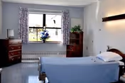 A simple nursing facility bedroom with a single bed with blue bedding, wooden dressers, a window with floral curtains and a potted plant.