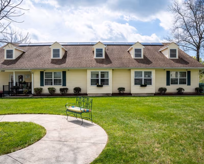 Exterior view of a single-story yellow elder care facility building with a brown roof and four dormer windows. There is a green lawn in front with a curved concrete walkway and a decorative metal bench with a cushion. Trees and a partly cloudy sky are visible in the background.