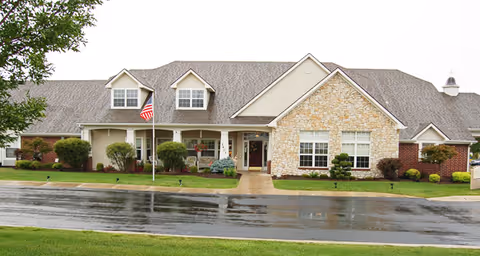 Exterior view of a single-story senior living facility building with a combination of stone and siding facade, multiple windows, a front porch with white columns, an American flag on a flagpole, and a wet driveway in front.