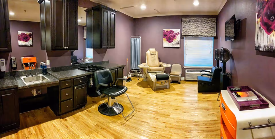 Interior of a salon or grooming room with purple walls and wooden flooring. The room features a black salon chair in front of a counter with a sink and cabinets, a beige reclining chair with armrests, a small beige chair, a black hair dryer chair, and a window with a patterned blind. There are floral paintings on the walls and various salon equipment and supplies visible.