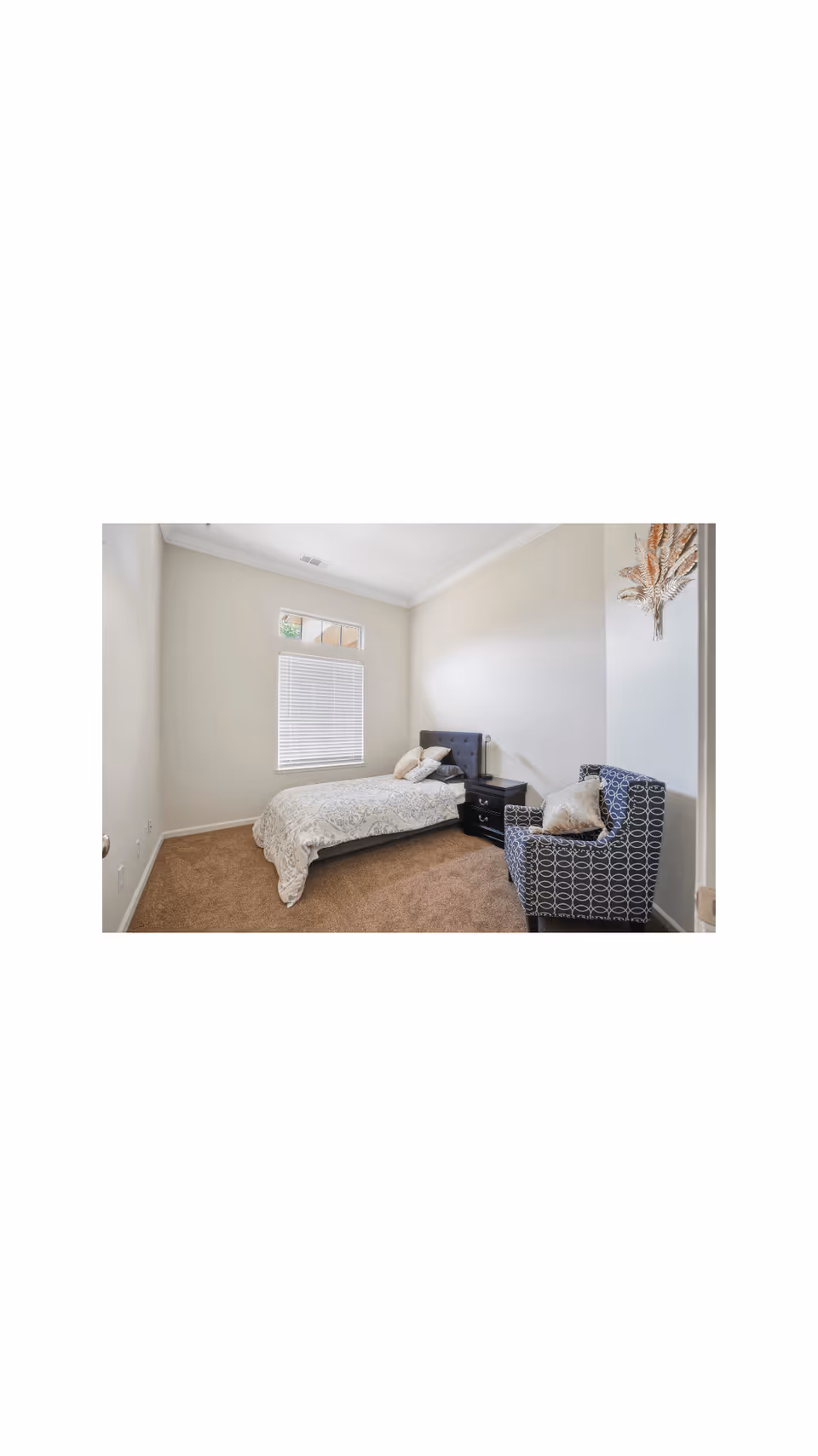 A small bedroom with a single bed against the wall, a nightstand with a lamp, a patterned armchair with a pillow, beige carpet, and a window with blinds letting in natural light.