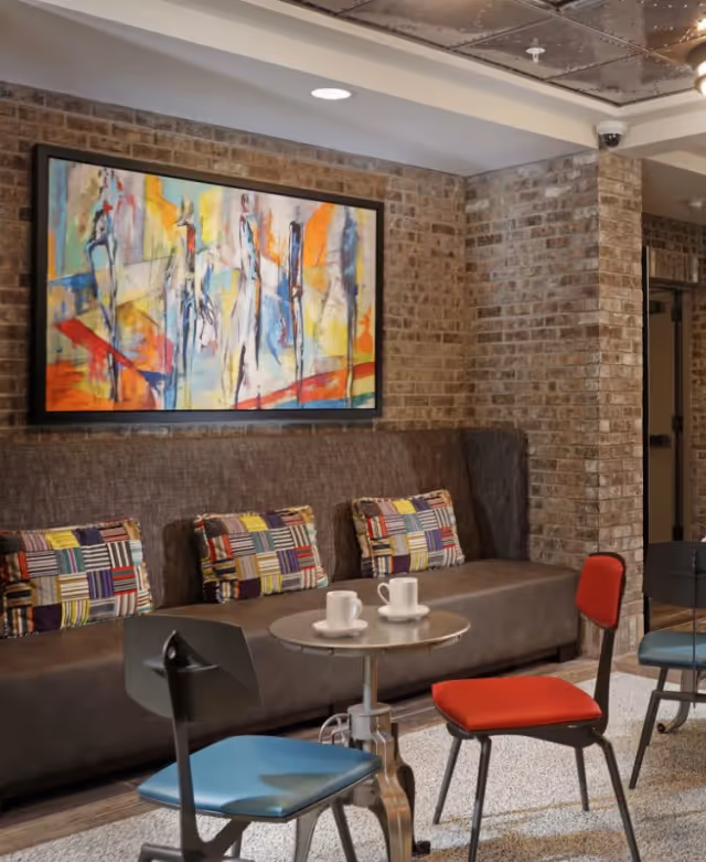 Lounge seating area with a brown upholstered bench, patterned throw pillows, colorful abstract painting, a small round table with two cups, and mixed-color chairs against an exposed brick wall.