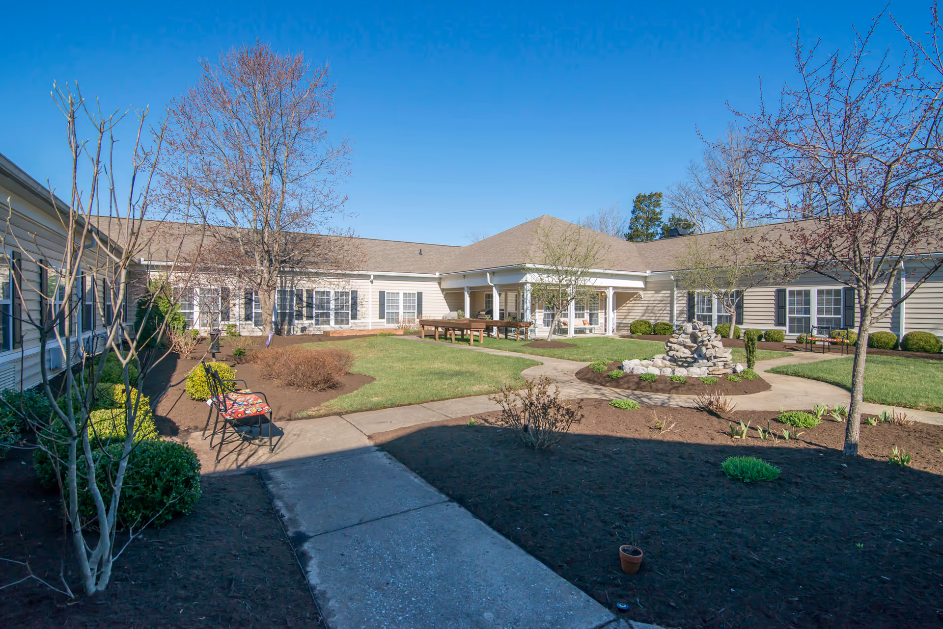 Outdoor courtyard area of a senior living facility with a paved walkway, landscaped garden beds, leafless trees, green grass, benches, and a small rock fountain in the center. The building surrounds the courtyard with beige siding and multiple windows under a clear blue sky.