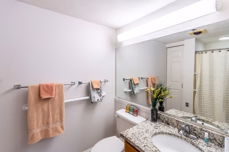 Bright bathroom showing a granite countertop vanity with sink and mirror, toilet, towel racks, and a shower with curtain.