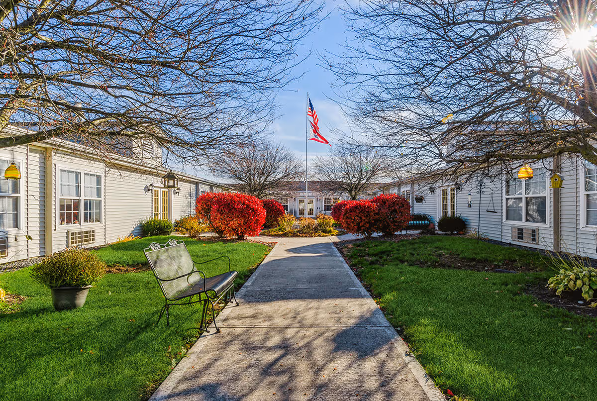 Outdoor courtyard area of a senior living facility with a concrete walkway leading to a building entrance. There are green lawns, red bushes, leafless trees, a bench on the left side, and an American flag flying on a flagpole in the center. The sky is clear and sunny.