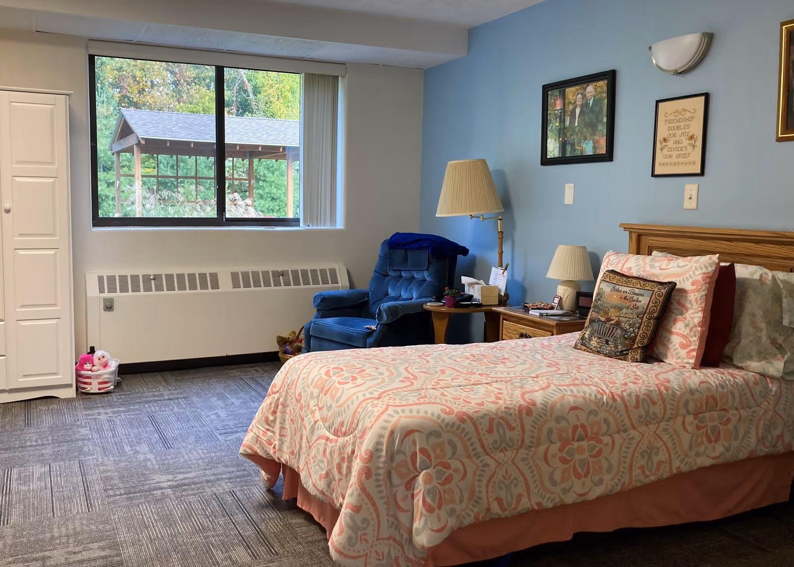 A cozy bedroom in a senior living facility with a single bed covered in a patterned pink and white bedspread. Next to the bed is a wooden nightstand with a lamp and some personal items. A blue upholstered armchair is placed near a window that shows a view of a wooden gazebo and greenery outside. The walls are painted light blue and decorated with framed pictures and a quote. A white closet door and a basket with stuffed animals are also visible.