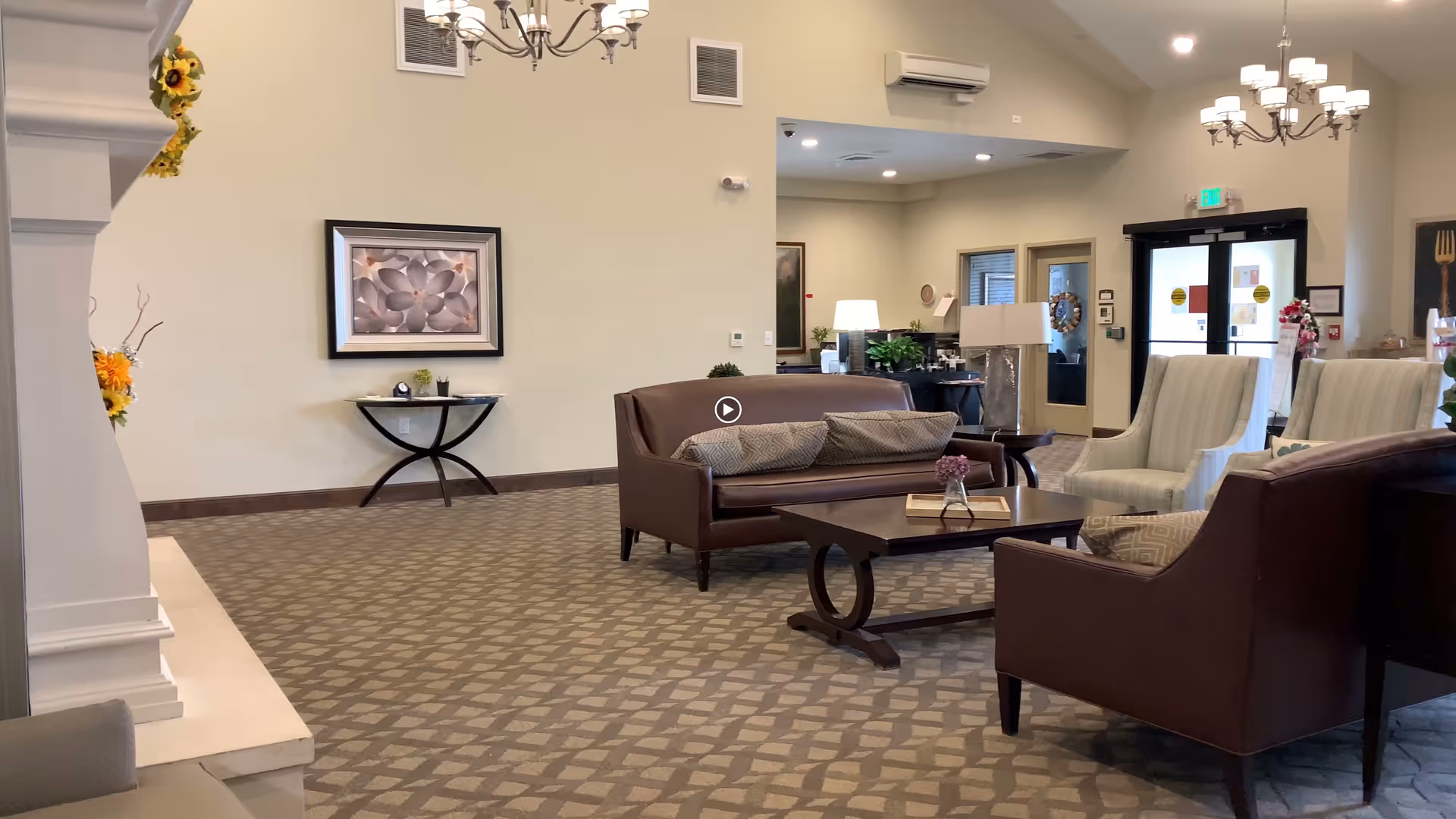 A spacious and well-lit assisted living facility common area with a patterned carpet, a brown leather sofa with cushions, two beige armchairs, a dark wooden coffee table with a small flower vase and a book, a side table with a lamp, a framed floral artwork on the wall, and a chandelier hanging from the ceiling. The entrance door and reception desk area are visible in the background.
