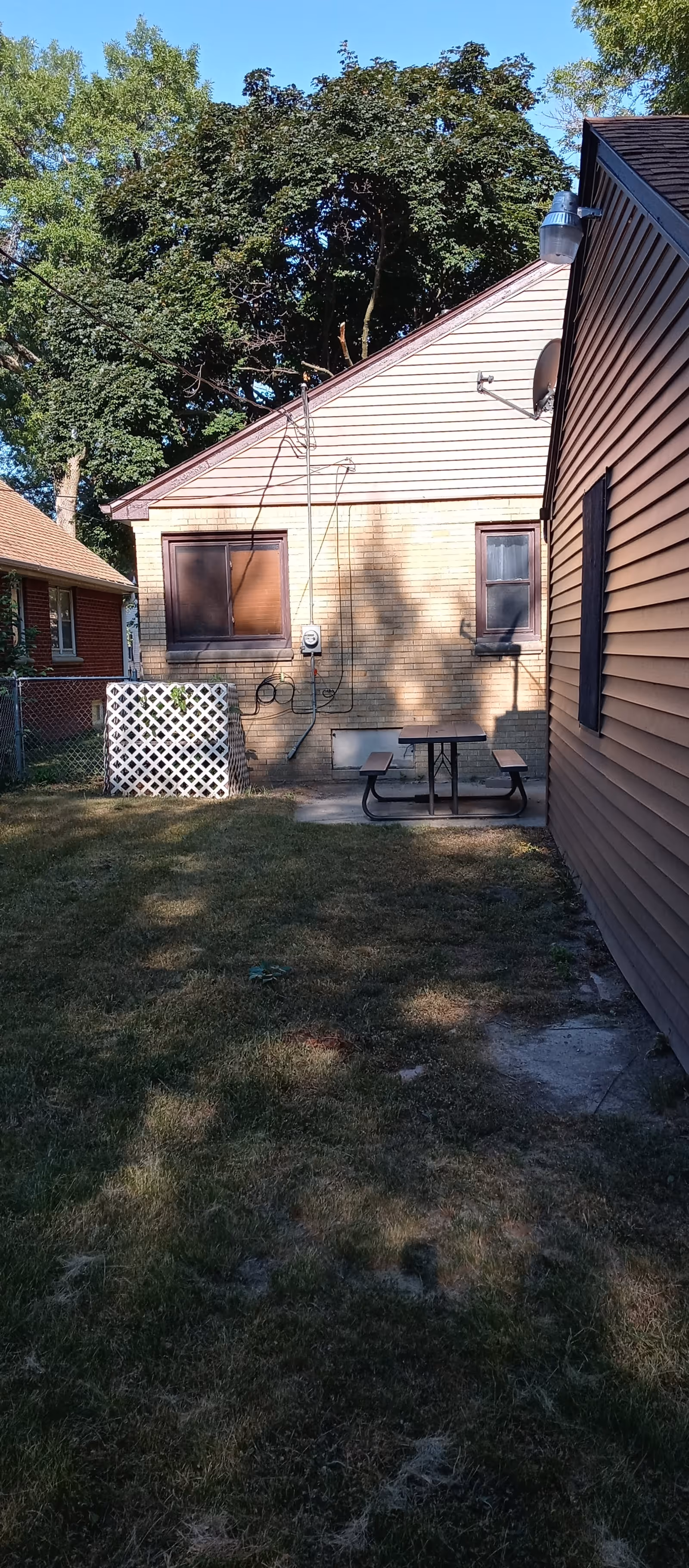 A small backyard area between two houses with grass and a concrete path. One house has beige siding and two windows, with a picnic table and benches placed against the wall. There are trees in the background under a clear blue sky.