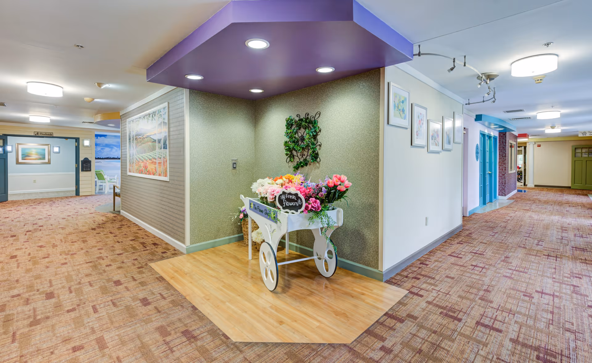 A brightly lit hallway in a senior living facility with patterned carpet and light-colored walls. In the center, there is a decorative white cart filled with colorful artificial flowers under a purple ceiling accent. The hallway features framed artwork on the walls and multiple doorways leading to other rooms.