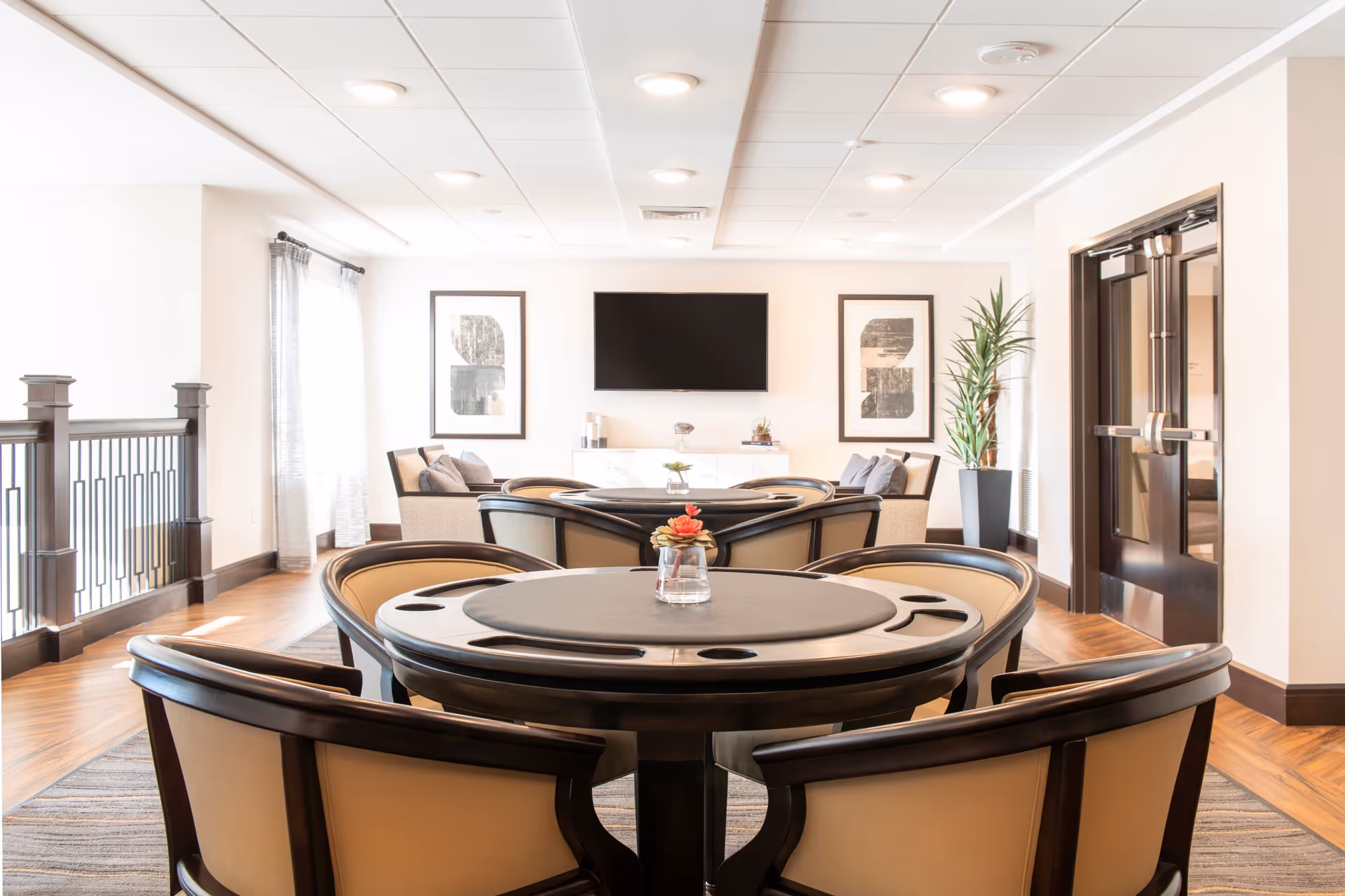 A bright and modern common area with round tables and cushioned chairs arranged for socializing or games. The room features a flat-screen TV mounted on the wall, two framed abstract artworks, a potted plant, and large windows with sheer curtains allowing natural light to fill the space.