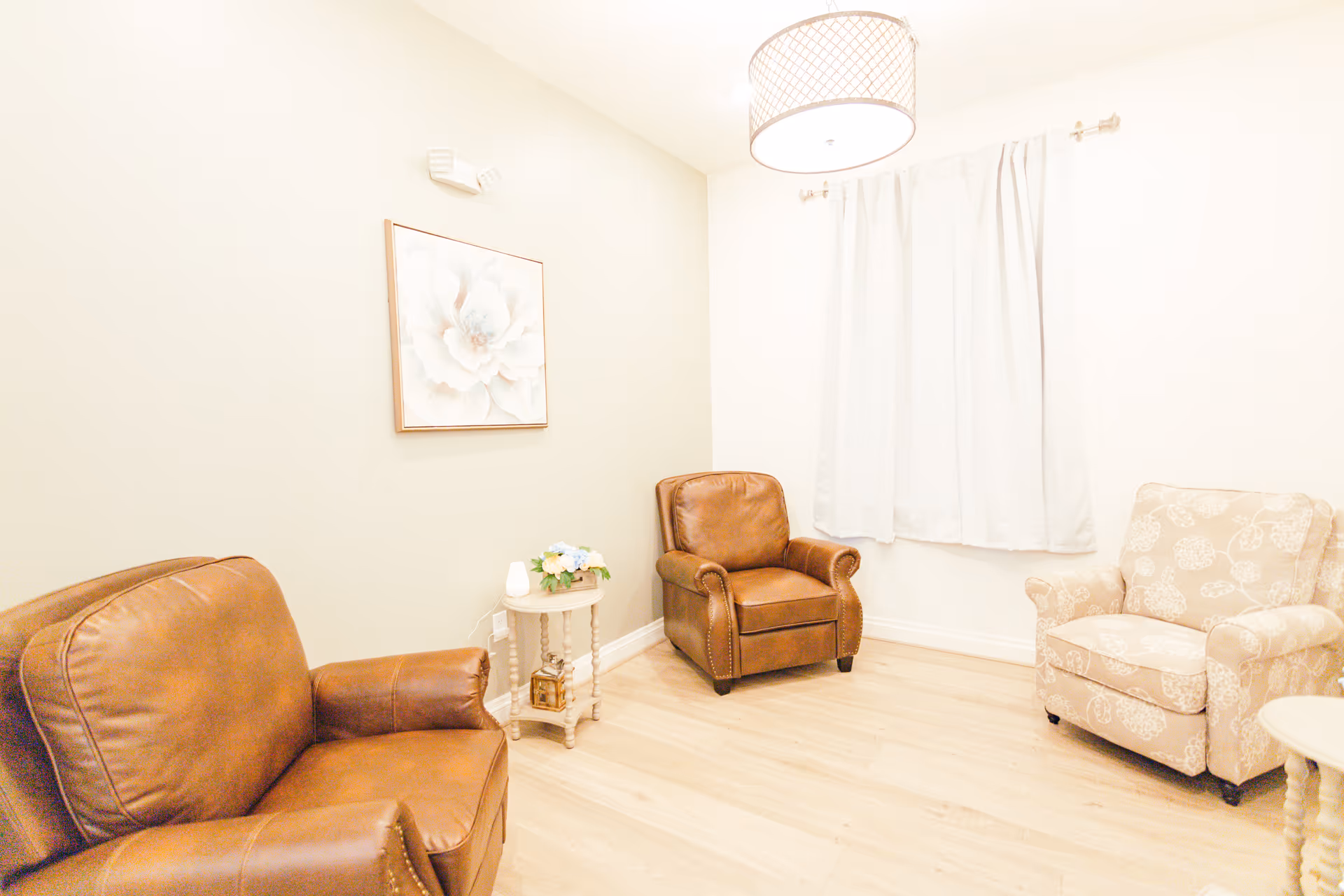 A cozy sitting area with two brown leather armchairs and one beige patterned armchair arranged around two small round side tables. The room has light-colored walls, a large floral painting, a window with white curtains, and a ceiling light fixture.