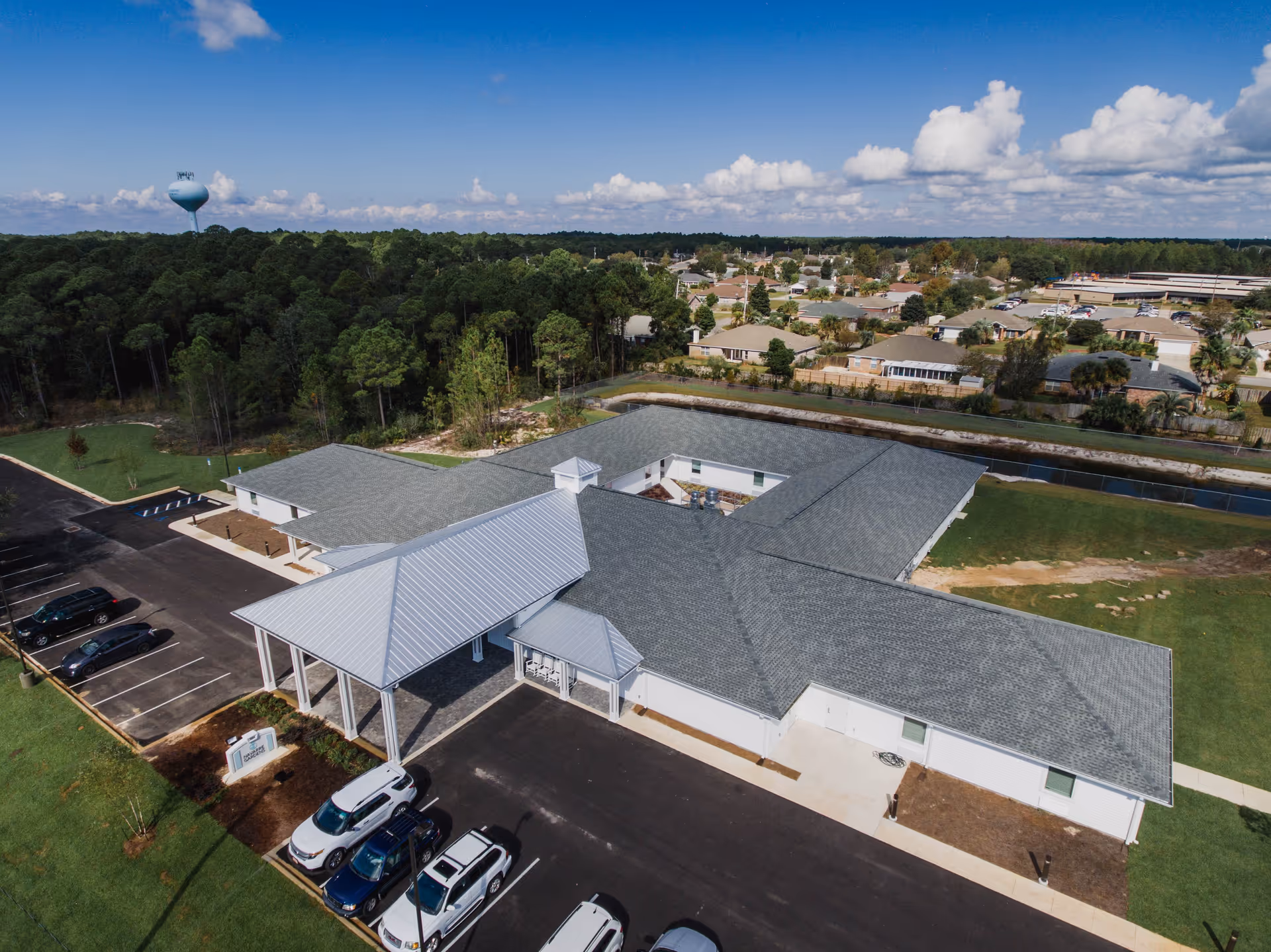 Aerial view of Navarre Gardens facility showing a single-story building with a gray roof and a covered entrance. Several cars are parked in the parking lot adjacent to the building. Surrounding the facility are green lawns, trees, and a residential neighborhood in the background under a partly cloudy blue sky.