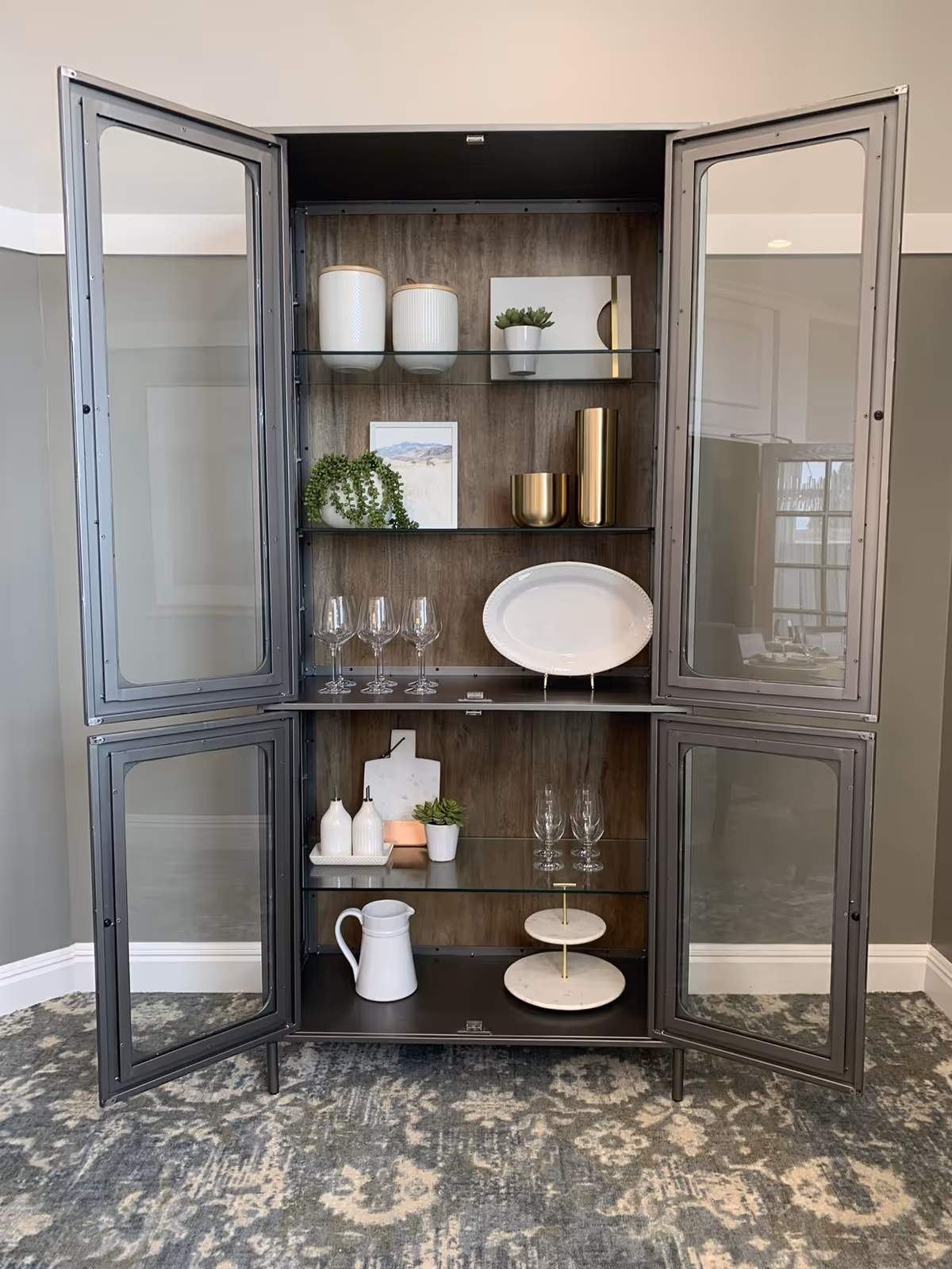 Glass-front display cabinet with shelves holding plates, wine glasses, pitchers, decorative vases and small potted plants in an interior space.