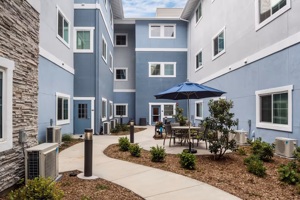 Courtyard with a paved walkway, patio table and umbrella, small shrubs, and a surrounding blue three-story building.