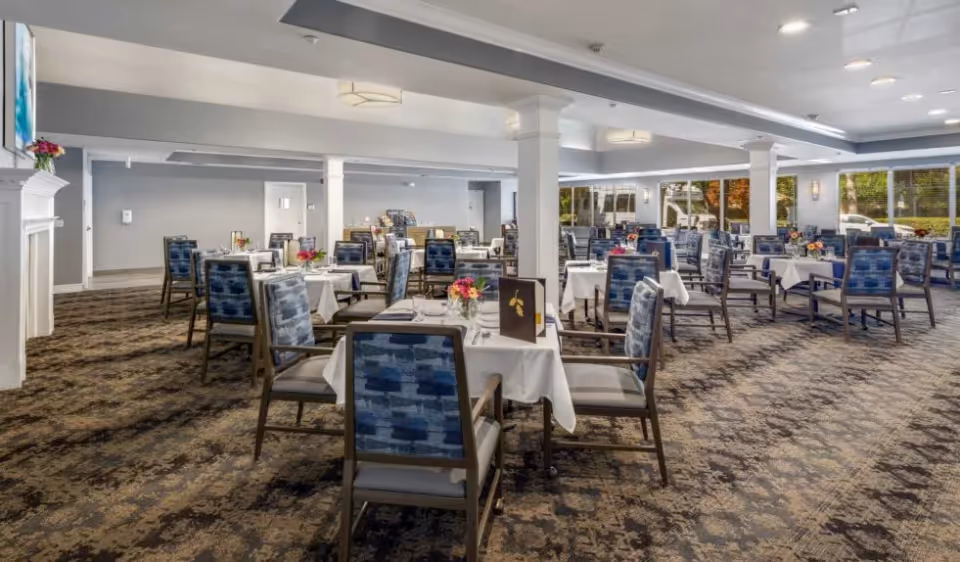 Spacious dining room with rows of tables covered in white tablecloths and blue-patterned chairs, each table set with flowers.