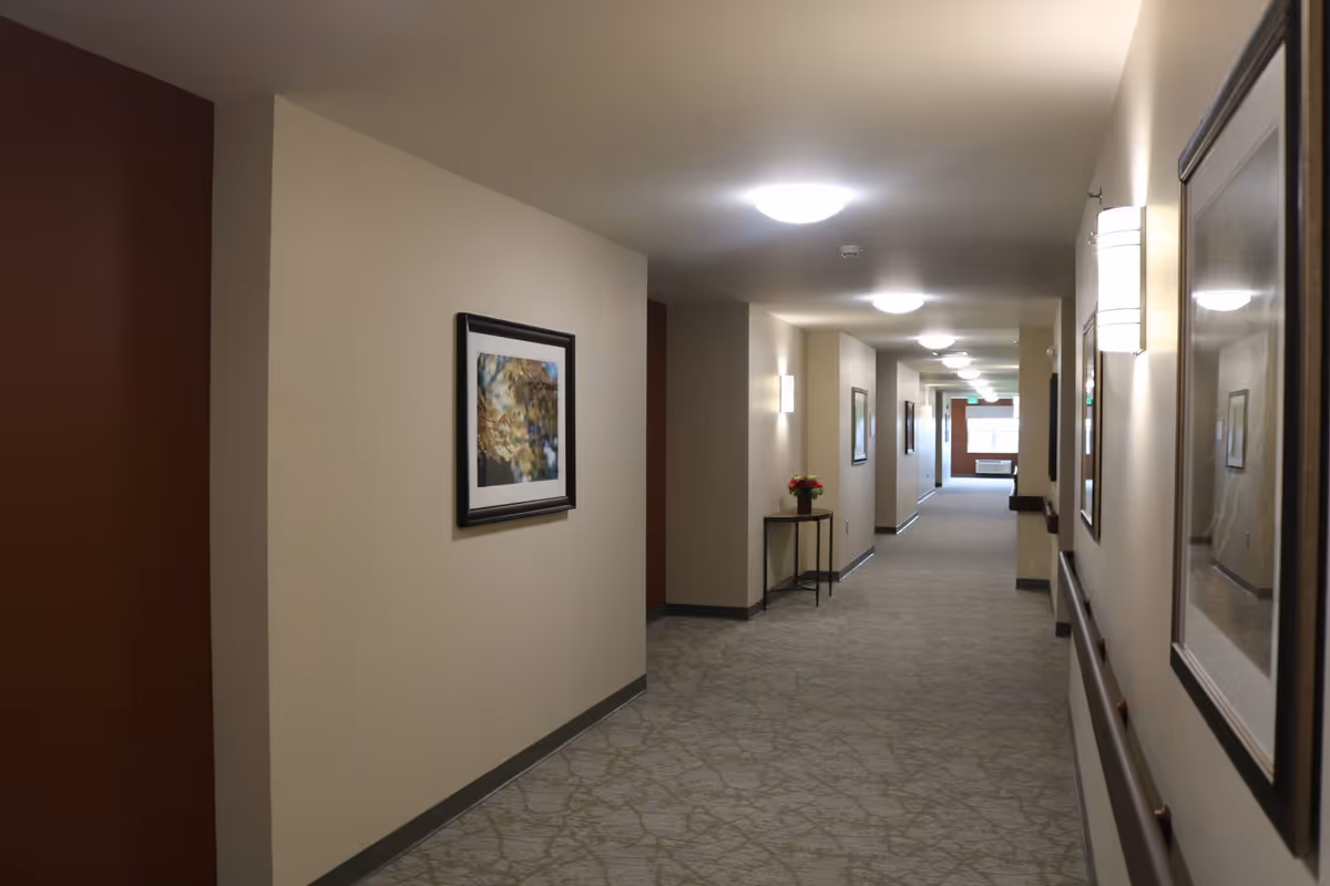 A well-lit interior hallway with framed artwork, wall sconces, handrails, and a small table with flowers.