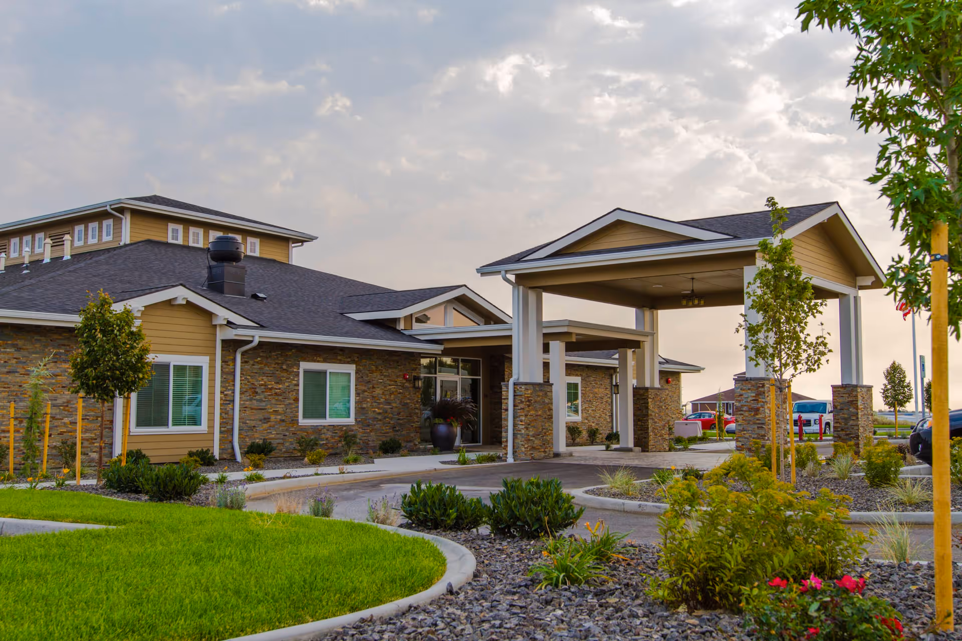 Exterior view of a senior living facility building with a covered entrance supported by stone pillars, surrounded by landscaped greenery, small trees, and a driveway under a partly cloudy sky.