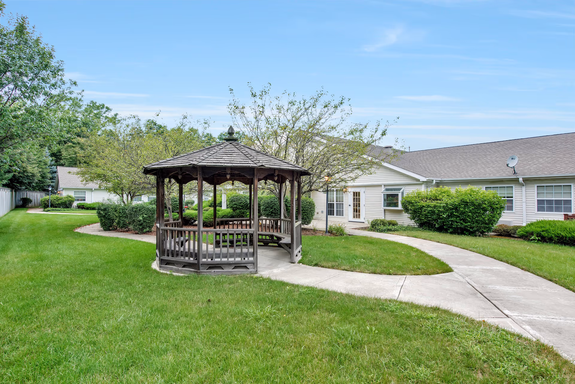 Outdoor area of Charter Senior Living of Hazel Crest featuring a wooden gazebo with benches on a concrete path surrounded by green grass, bushes, and trees, with a single-story building in the background under a blue sky.