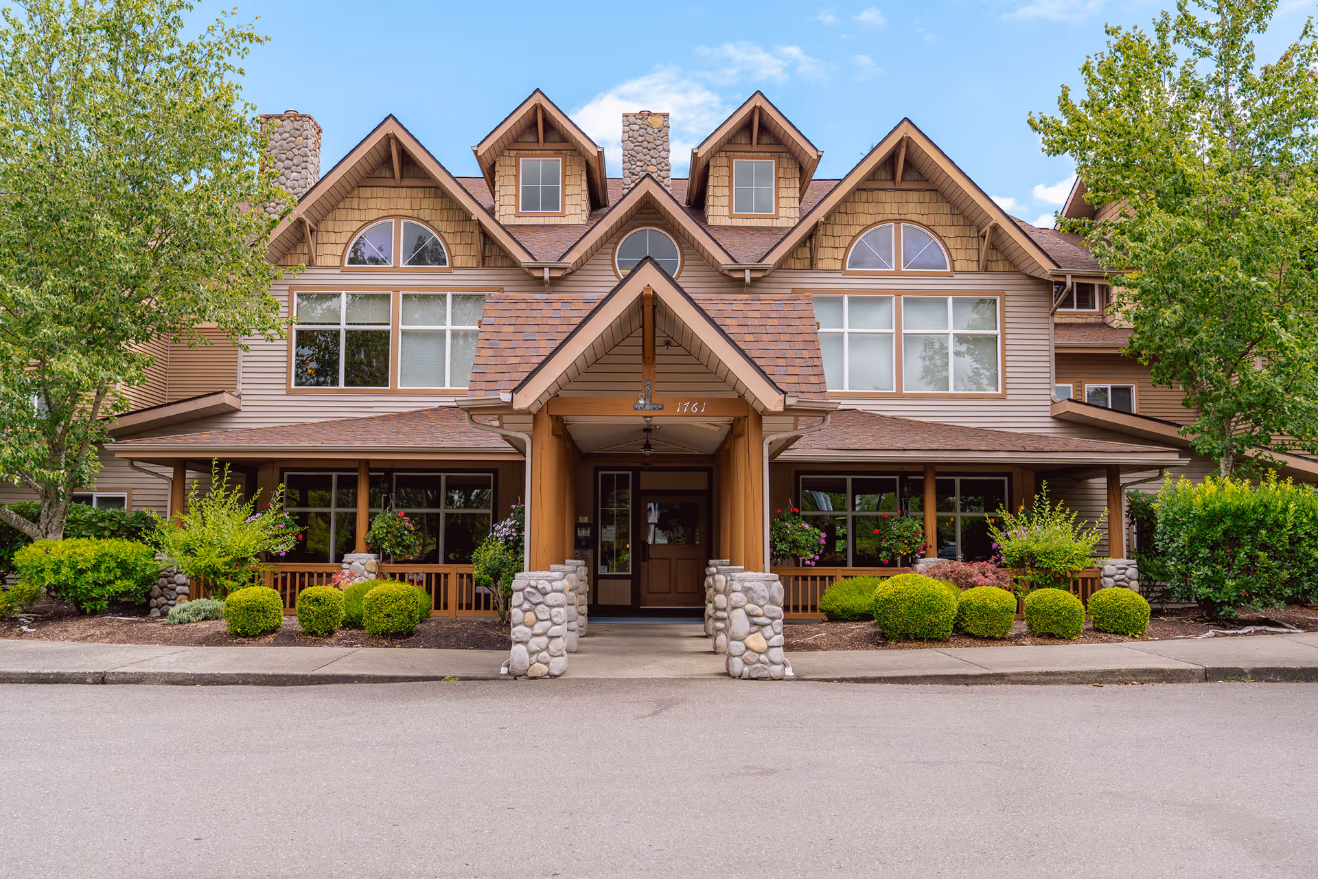 Front exterior view of a senior living facility named Vineyard Park of Port Orchard, featuring a large building with multiple windows, stone pillars, a covered entrance, and well-maintained landscaping with bushes and trees.