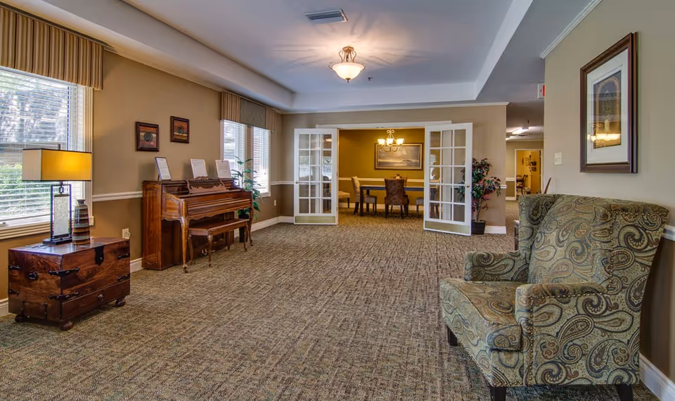 A cozy interior common area with patterned carpet, a wooden piano with a bench, a side table with a lamp, and a comfortable upholstered armchair. The room has large windows with blinds and valances, and French doors leading to a dining area with a table and chairs under a chandelier. The walls are decorated with framed artwork and plants are placed near the doors.