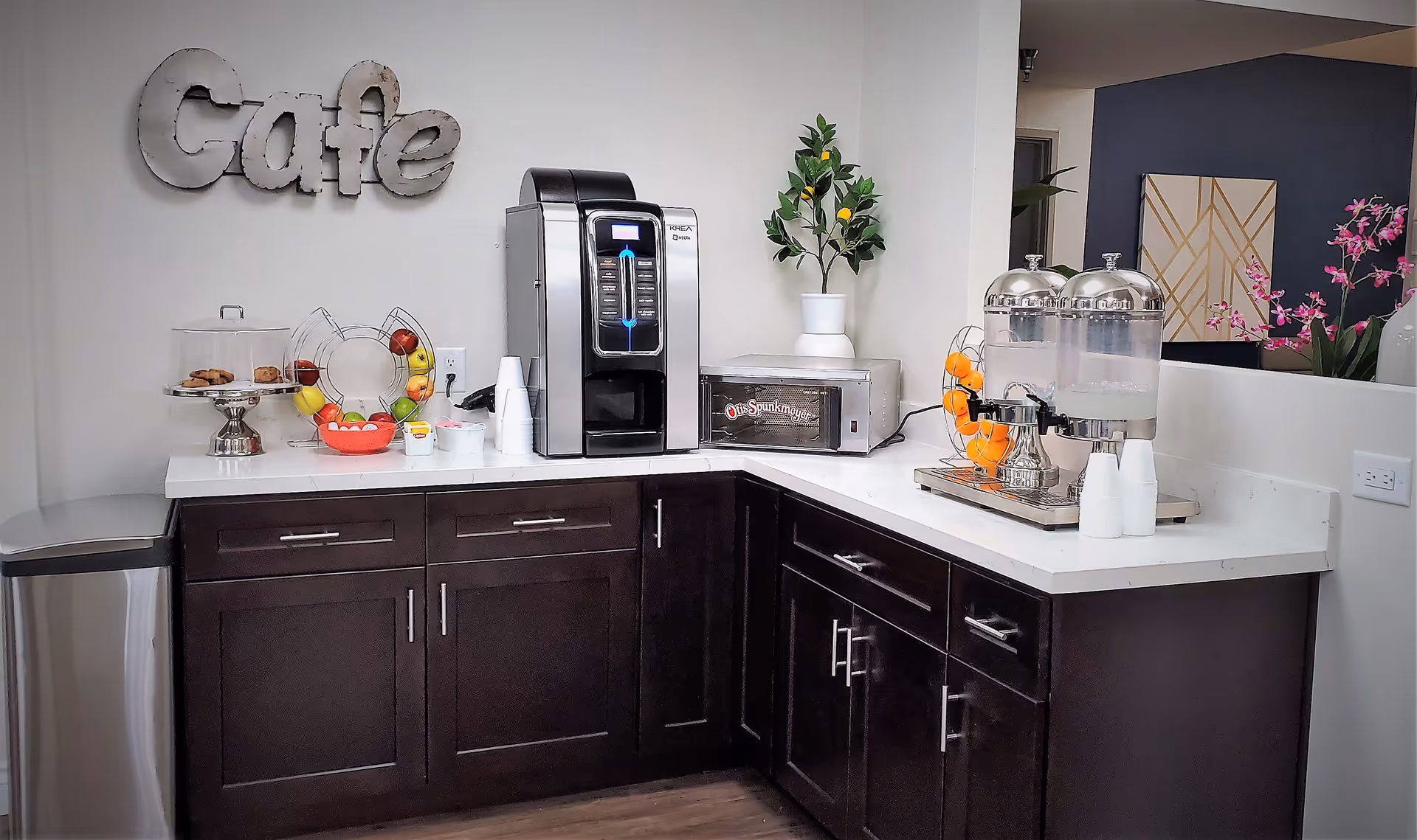 A small cafe area with dark wood cabinets and white countertops. On the counter, there is a coffee machine, a toaster oven, a fruit basket with apples and bananas, a container with cookies, a water dispenser with cups, and a decorative plant. The word 'Cafe' is mounted on the wall above the counter.