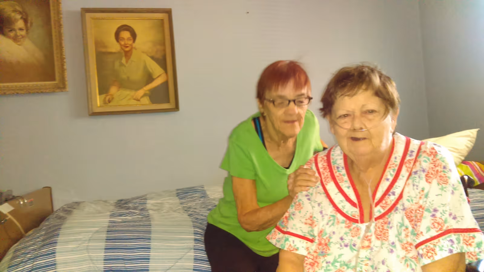 Two elderly women in a bedroom. One woman with short red hair and glasses wearing a green shirt is standing behind another woman with short brown hair wearing a floral nightgown and an oxygen tube. The bedroom has a bed with a striped blue and white bedspread and two framed portraits on the wall.