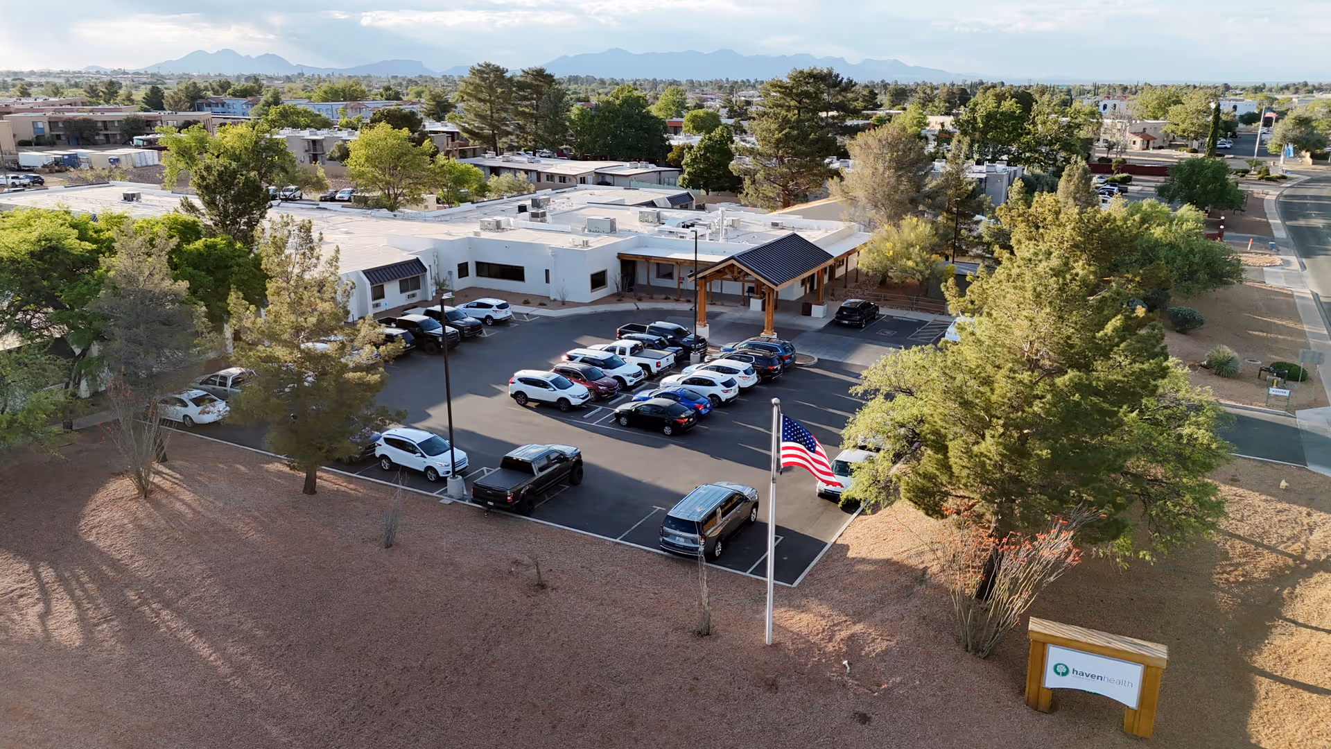 Aerial view of Haven Health Sierra Vista facility showing a single-story building with a parking lot filled with cars, surrounded by trees and landscaping. An American flag is visible near the entrance, and a sign with the Haven Health logo is positioned on the lawn in front of the building. Mountains can be seen in the background under a partly cloudy sky.