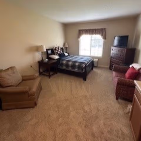 A bedroom with a bed covered in a checkered blanket, two bedside tables with lamps, a window with a valance, a dresser with a TV on top, a brown armchair, and a red upholstered chair.