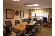A conference room with a long rectangular table surrounded by black office chairs. The table has a decorative centerpiece and some items on it. The room has beige walls with framed artwork and a decorative sunburst wall piece. A large window with a striped valance lets in natural light, and there are two desks with chairs along the window wall.
