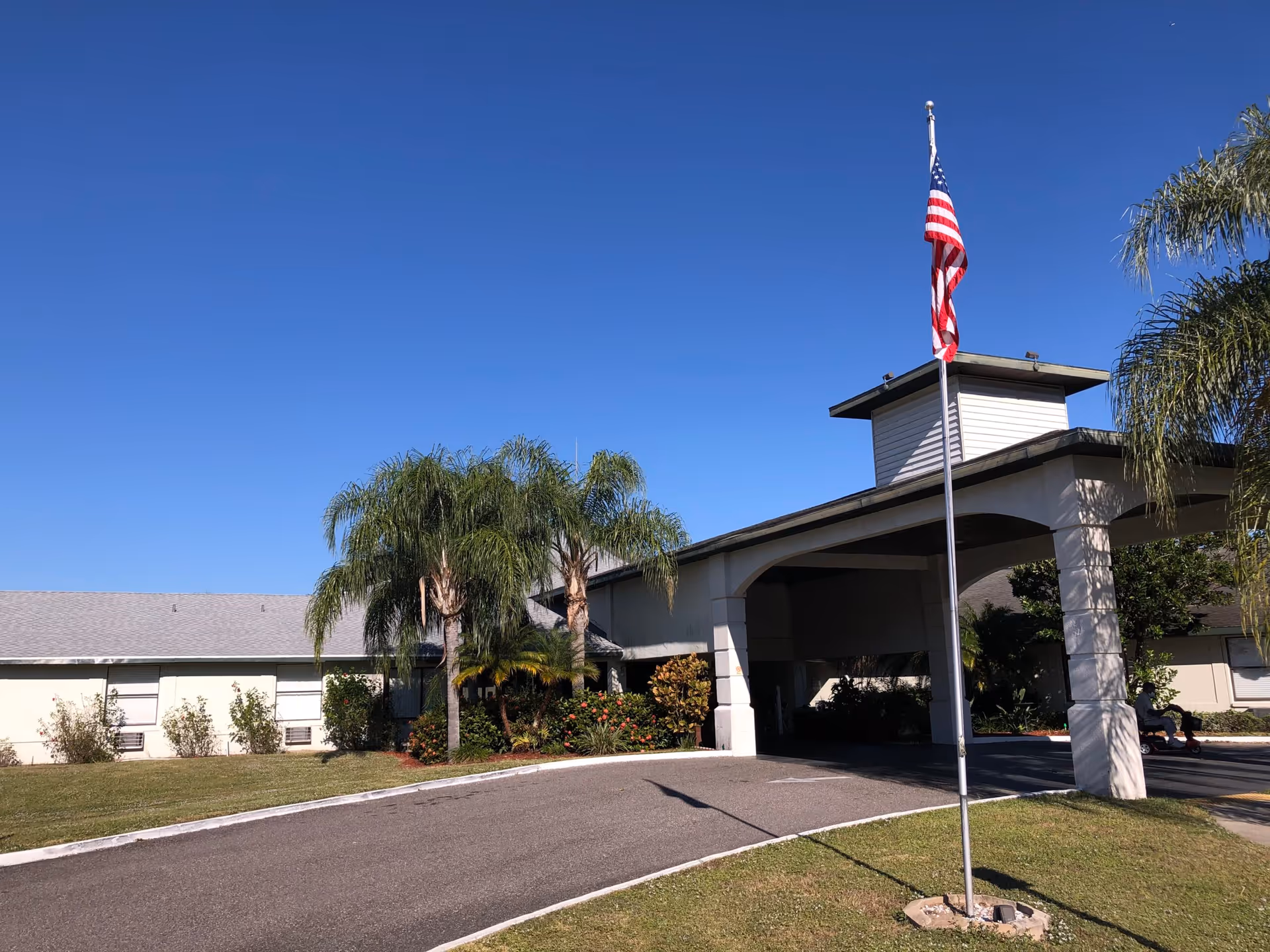 Exterior view of Sunny Hills Alf - Assisted Living Facility Sebring showing the front entrance with a covered driveway, an American flag on a flagpole, palm trees, and a clear blue sky.
