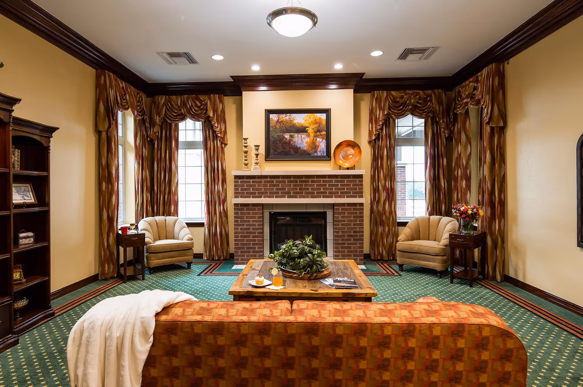 A cozy living room with a patterned orange sofa draped with a white blanket, a wooden coffee table with a plant and drinks, two beige armchairs beside small wooden side tables, a brick fireplace with decorative items on the mantel, large windows with patterned curtains, and a wooden bookshelf on the left.