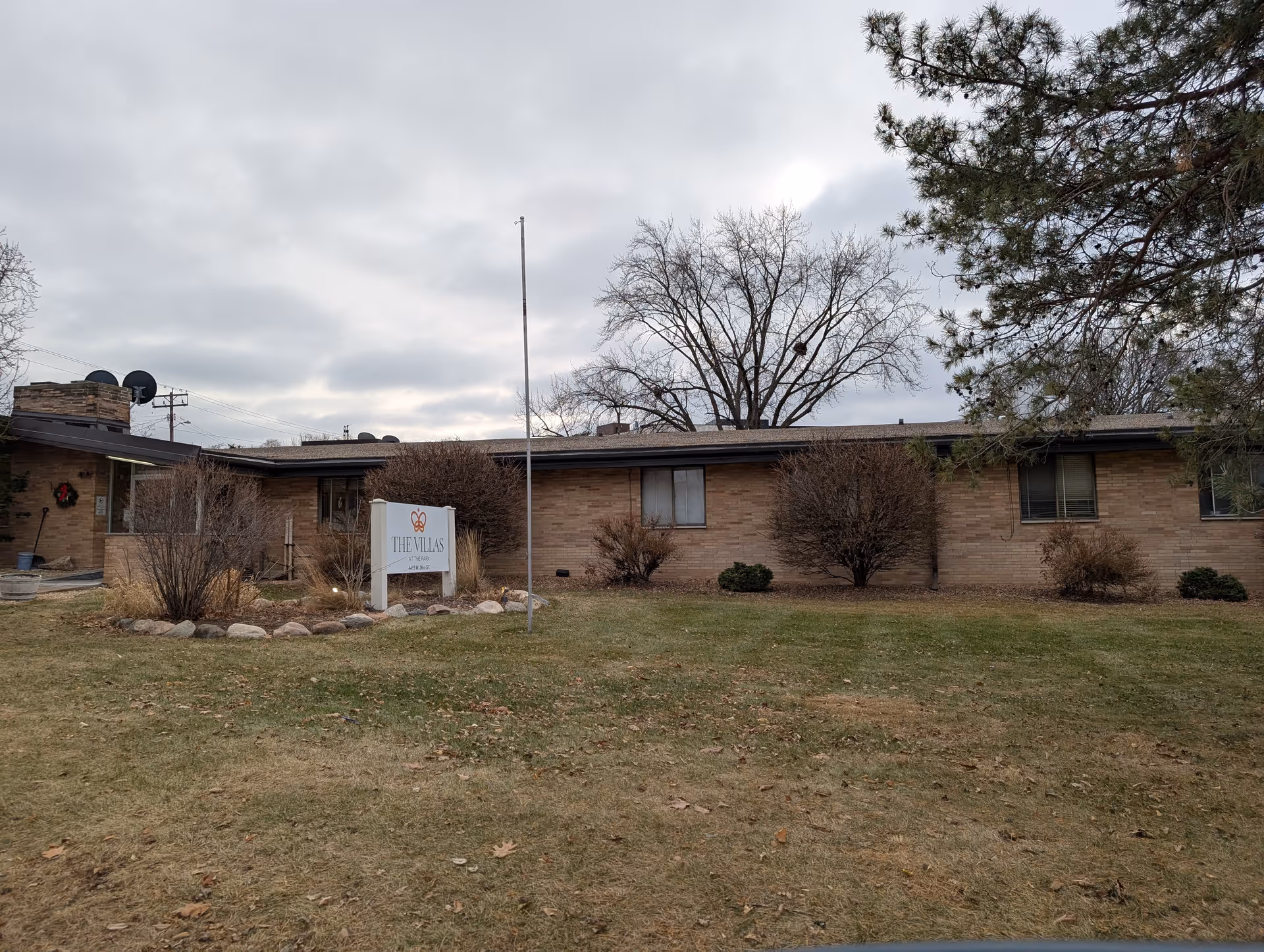 Single-story brick senior living building with a lawn, shrubs, and a freestanding sign in front under a cloudy sky.