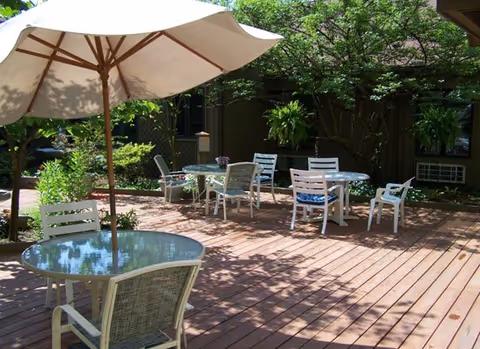 Outdoor patio area with wooden deck flooring, several white tables and chairs, and a large beige umbrella providing shade. Green plants and trees surround the space, creating a peaceful garden atmosphere.