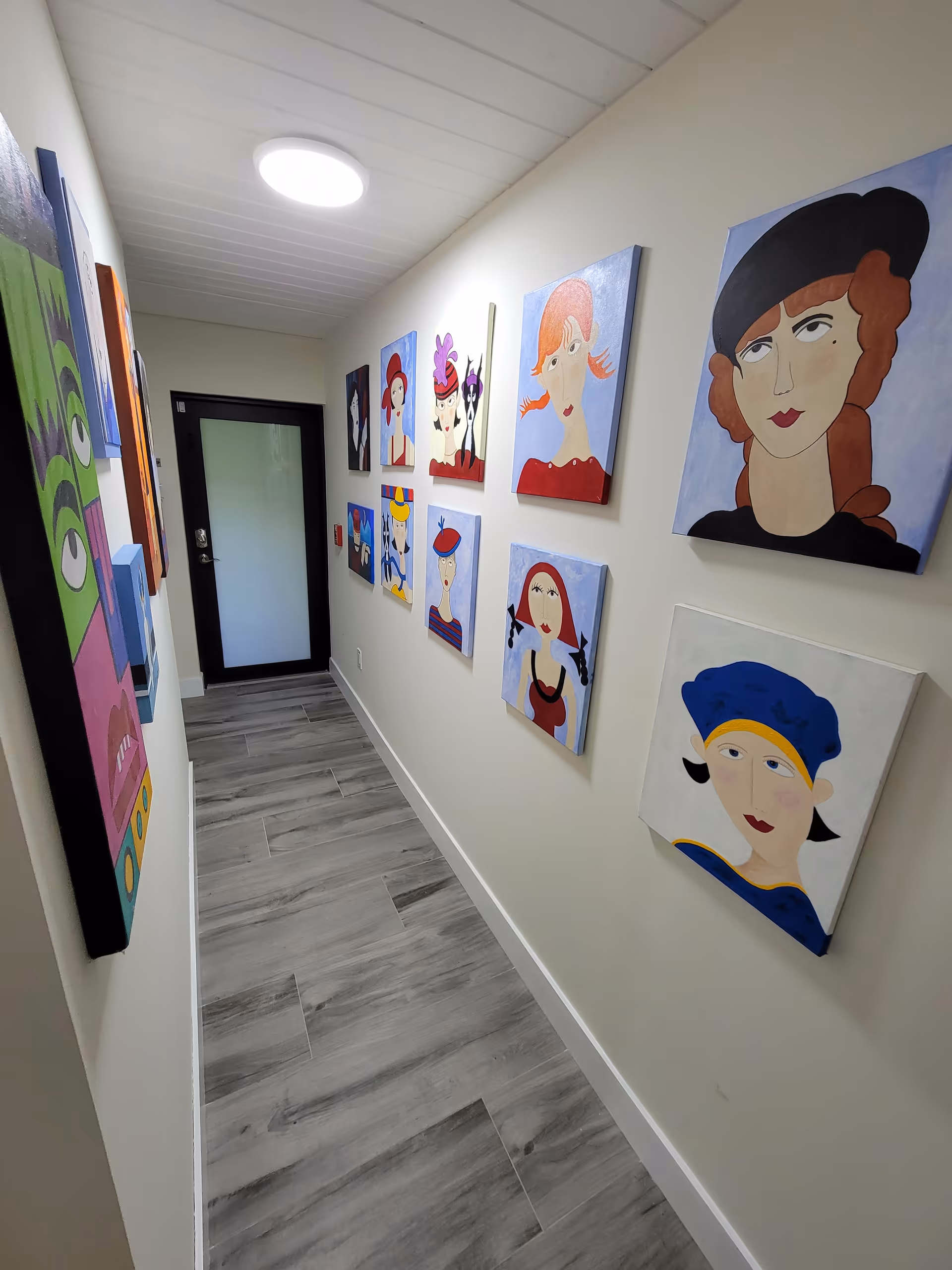 A narrow hallway with light-colored walls and gray wood-patterned floor tiles. The walls are decorated with colorful, abstract portraits of women wearing hats and various outfits. At the end of the hallway is a door with a frosted glass panel and a black frame. A round ceiling light illuminates the space.