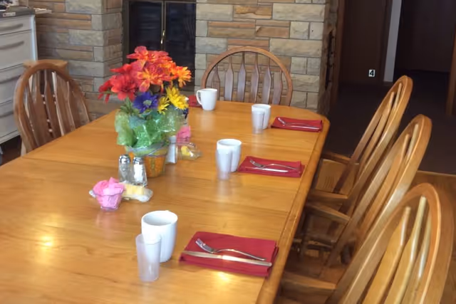 A wooden dining table set with red napkins, silverware, white cups, and a colorful flower arrangement in the center. The background features a stone fireplace and wooden chairs around the table.