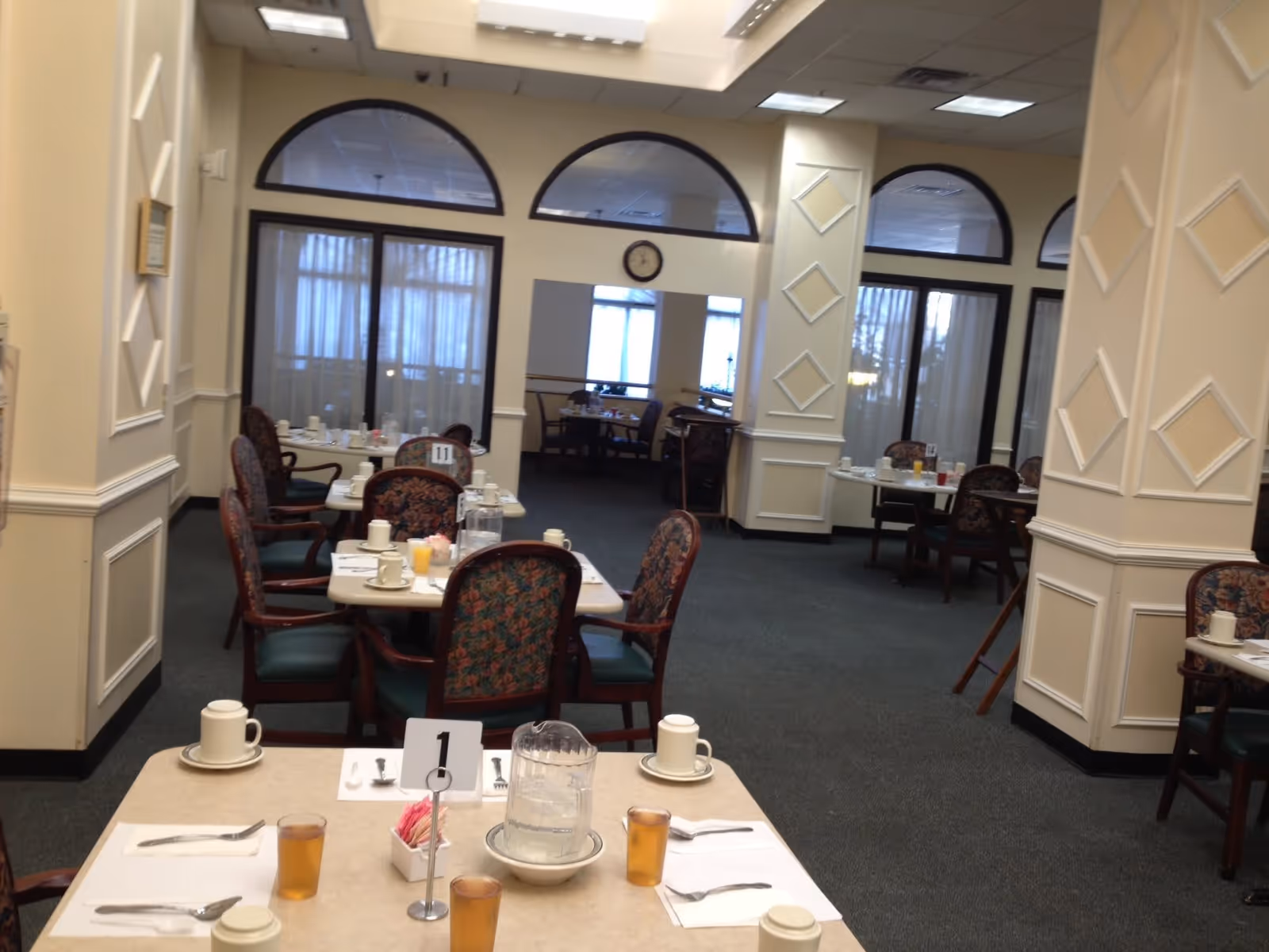 Interior view of a dining room in an assisted living facility with multiple tables set for meals. Each table has chairs with floral upholstery, place settings including cups, glasses with iced tea, utensils, and pitchers of water. The room features cream-colored walls with decorative molding and large windows with sheer curtains.