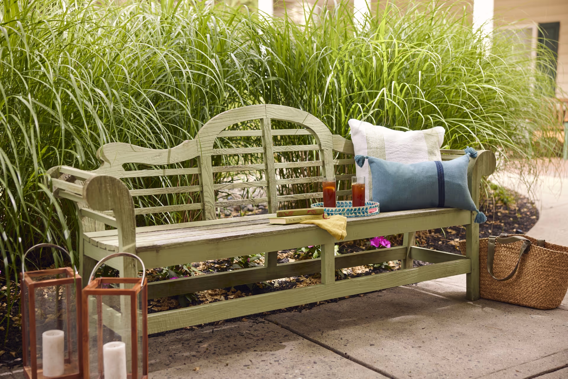 A light green wooden bench outdoors with two decorative pillows, a tray holding two glasses of iced tea, and a book with a yellow cloth on the bench. Next to the bench are two lanterns with white candles inside and a woven basket bag on the ground. Tall green grass plants are in the background.