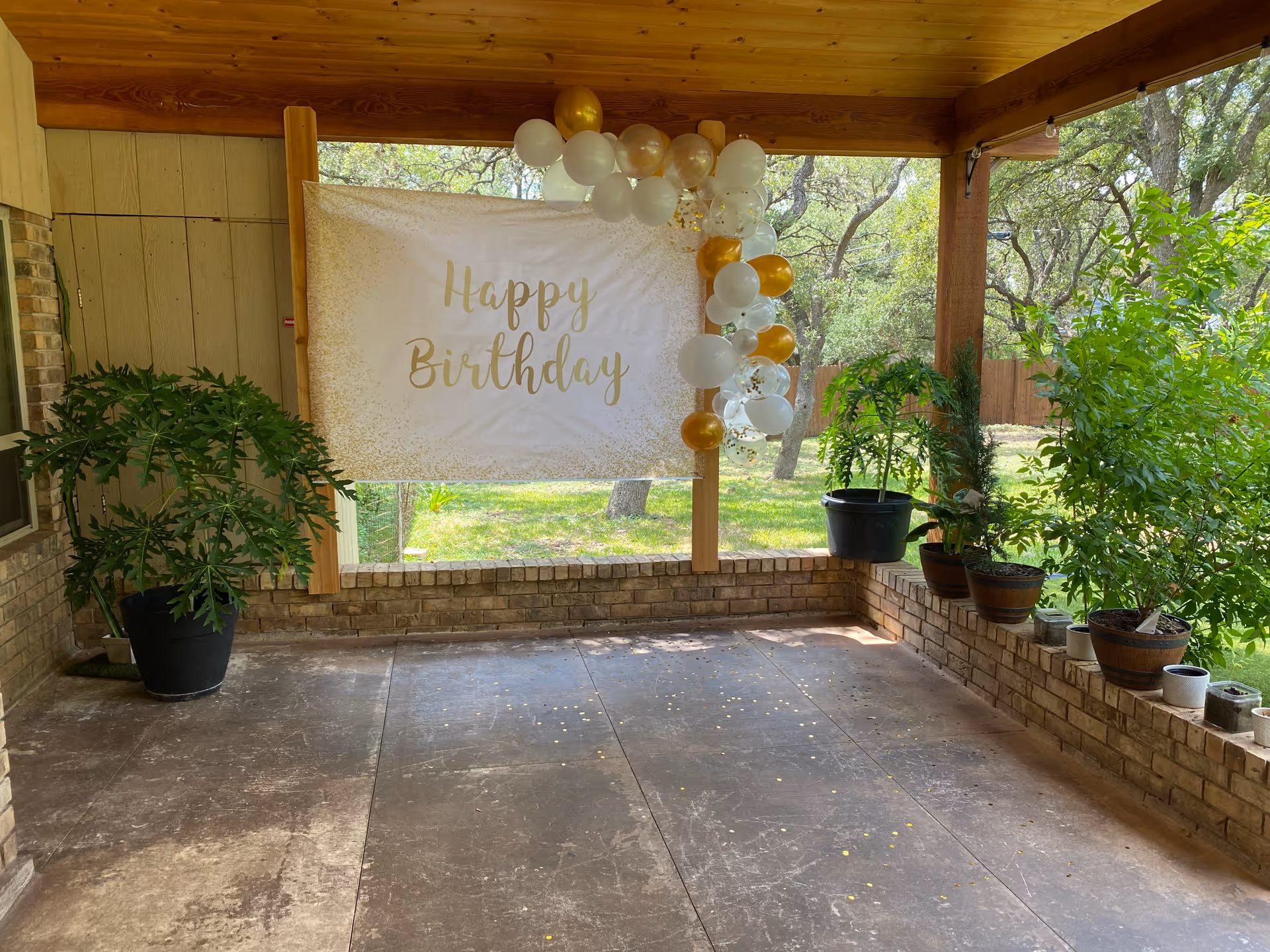 Covered outdoor patio area decorated with a white and gold 'Happy Birthday' banner and matching balloons. Several potted plants are placed along the brick ledge and floor. Trees and greenery are visible in the background.