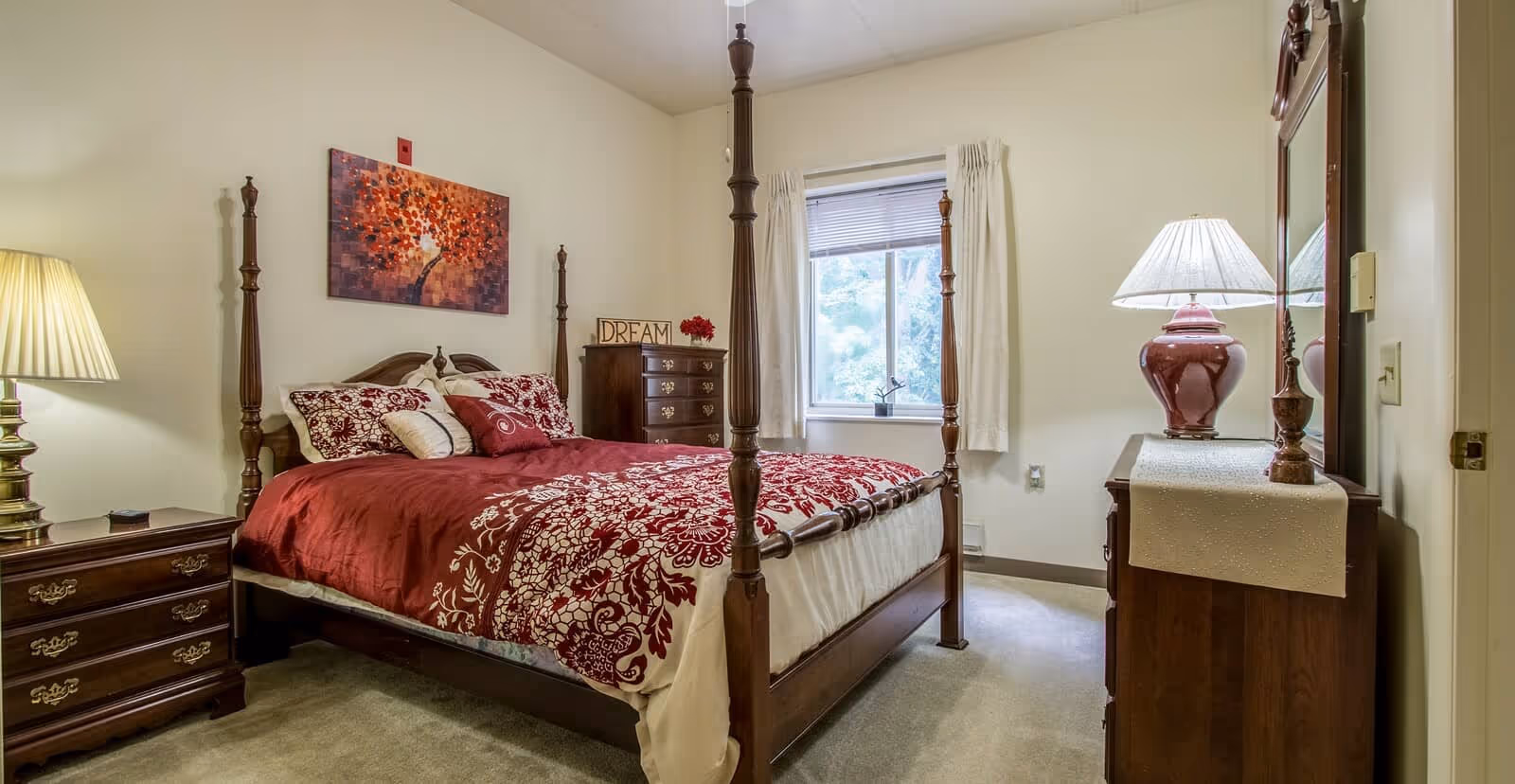 A cozy bedroom with a wooden four-poster bed dressed in red and white floral bedding. There are two wooden dressers, one with a lamp and the other with a decorative sign that says 'DREAM' and a small flower arrangement. A window with white curtains lets in natural light, and a colorful painting of a tree hangs on the wall above the bed.