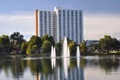 A tall white multi-story building named Westminster Towers is seen behind a lake with three water fountains. Trees and greenery surround the lake, and the sky is partly cloudy.
