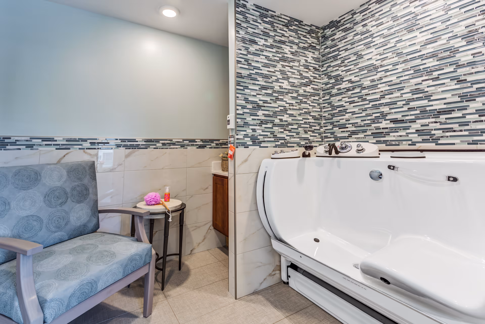 A bathroom area featuring a white walk-in bathtub with multiple control knobs and a built-in seat. The walls are decorated with a mosaic tile pattern in shades of blue, gray, and white. Next to the bathtub is a small round table holding a pink bath sponge and a bottle of soap or lotion. A cushioned chair with a circular pattern on the upholstery is positioned beside the table. The floor is tiled in a light gray color.